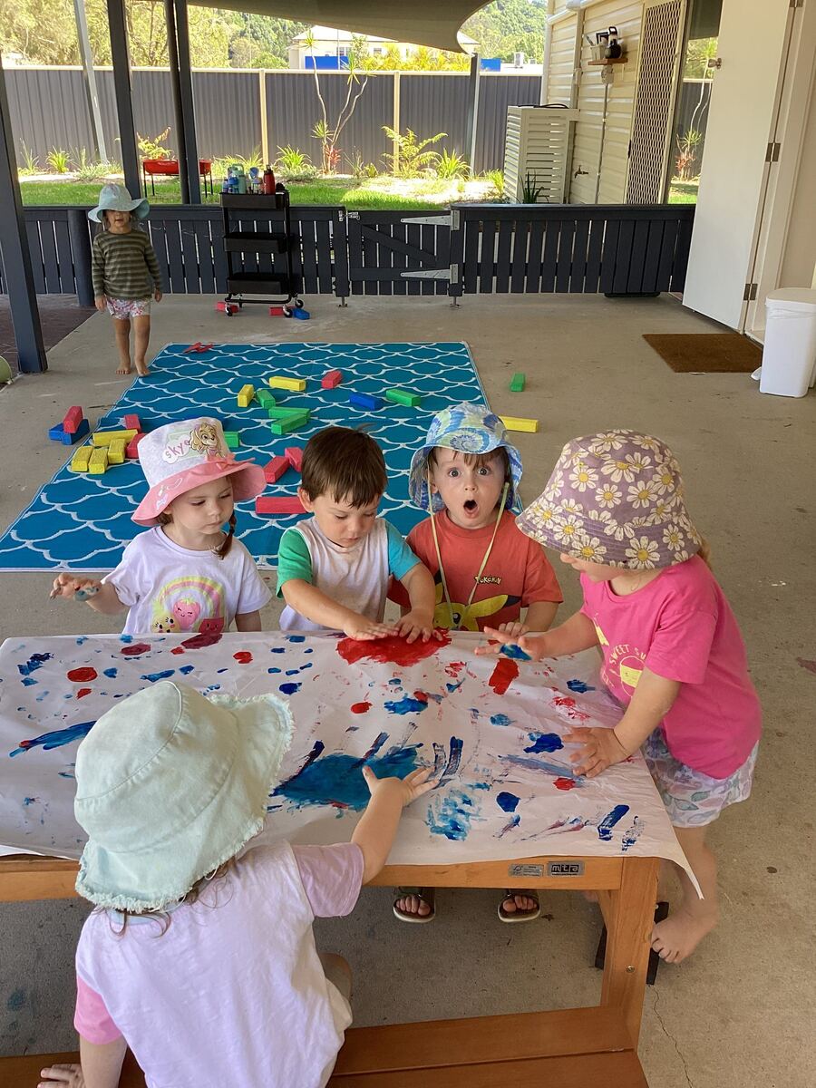 Children paint outdoors at a table, guided by an educator