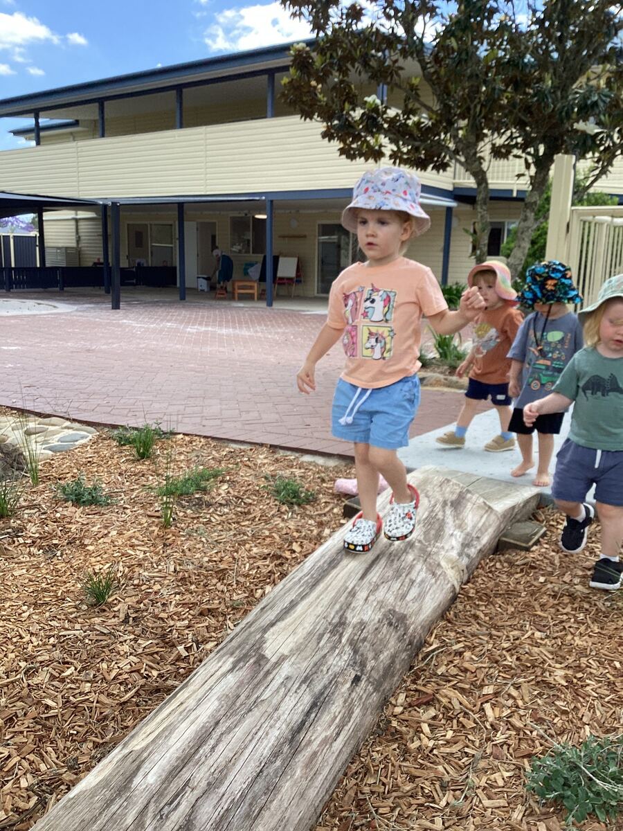 Children walk along a log in the outdoor play area, supervised by educators